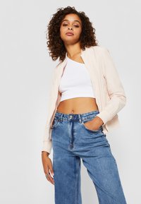 Světle růžová bomber jacket s zipovým uzávěrem, žebrovanými manžetami, nošená přes bílý crop top a modré džíny s vysokým pasem. Rámované kudrnaté vlasy.