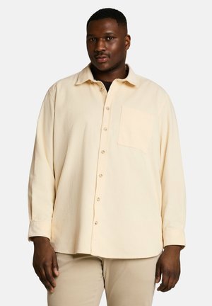 Mann trägt ein hellbeiges Hemd mit Knöpfen und beige Hose, steht vor einem schlichten weißen Hintergrund.