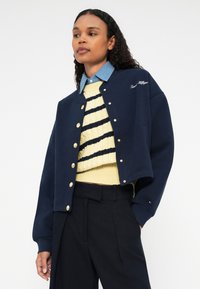 Námornícky modrá skrátená bunda so zlatými cvokmi, kombinovaná so žltým pruhovaným sveterom a námorníckymi nohavicami s vysokým pásom. Viditeľný je prevrátený golier džínsovej košele.