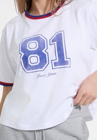 Valkoinen puuvillainen t-paita, jossa on punaiset ja siniset ribbi-kaula-aukko, ylisuhteellinen istuvuus ja suuret siniset numero 81 sekä "Guess Jeans" -logo painettuna alapuolelle.