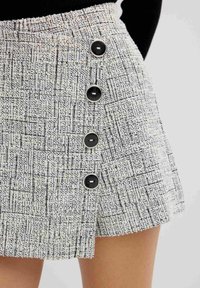 Minifalda blanca y negra texturizada con forma ligeramente acampanada, que cuenta con cuatro botones negros con acentos plateados en el lado.