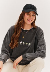 Tmavě šedá nadměrná mikina se zimními postavičkami na přední straně, doplněná černou baseballovou čepicí a barevnými náhrdelníky ve vrstvách.