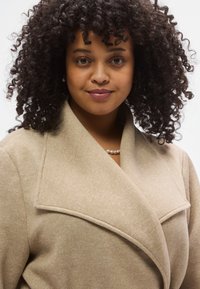 Smėlio spalvos apsiaustas paltas su plačiomis atlapomis, tekstūruotu audiniu ir švelniu blizgesiu. Turi apykaklę ir derinamas su perlų vėriniu.