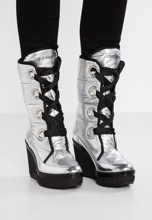 Glänzende silberne Keilstiefel mit schwarzen dicken Schnürsenkeln, schwarzen Sohlen und dem „Calvin Klein“ Logo auf gepolsterten Zungen, getragen zu schwarzen Hosen.