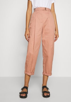 Personne portant un pantalon taille haute, court, rose poussiéreux avec des plis et des ourlets retroussés, associé à un t-shirt blanc et des sandales noires.