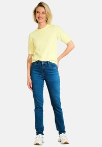 Żółty sweter krótki rękaw z okrągłym dekoltem, w połączeniu z niebieskimi dżinsami skinny i białymi butami sportowymi. Gładka tekstura, swobodny krój.