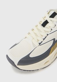White athletic shoe with a mesh upper, black and gold accents, thick sole, prominent laces, and a curved design for comfort.