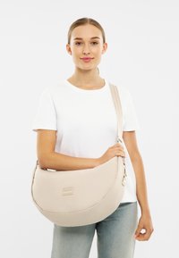Bežová ramenná taška v tvare polmesiaca, vyrobená z mäkkej textílie, s diskrétnym logom a nastaviteľným popruhom. Model nosí biele tričko.