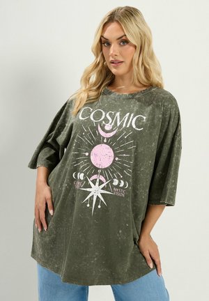 Frau mit langen blonden Haaren trägt ein übergroßes, ausgewaschenes grünes T-Shirt mit pinker kosmischer Sonne, Mond, Sternen und den Texten "COSMIC" und "Mystic Vision."
