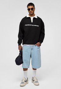 Zwarte sweatshirt met witte opdruk "PEACEFULNESS", witte kraag, gecombineerd met lichtblauwe denimshorts, witte sokken en beige sneakers.