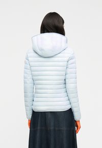 Γυναίκα που φοράει ανοιχτό μπλε κοντό μπουφάν με κουκούλα και σκουρόχρωμη φούστα από τζιν, εμφανίζεται από πίσω με φόντο ένα απλό λευκό.