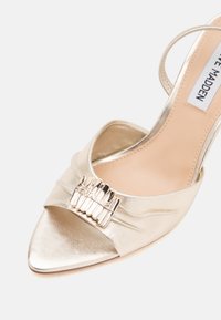 Χρυσό γυναικείο παπούτσι slingback με μυτερή μύτη, διακοσμητική χρυσή αγκράφα στο μπροστινό μέρος και ορατή ετικέτα Steve Madden στην εσωτερική σόλα.