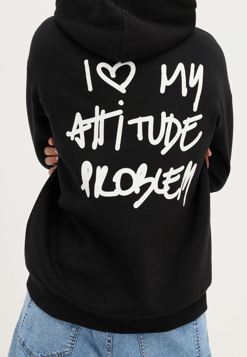 Personne portant un sweat à capuche noir avec un texte blanc style graffiti disant "J'aime mon problème d'attitude" dans le dos, associé à un jean bleu.