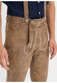 Lederhosen marroni con bretelle, caratterizzati da motivi floreali ricamati e cuciture decorative. Tessuto testurizzato con due tasche laterali.