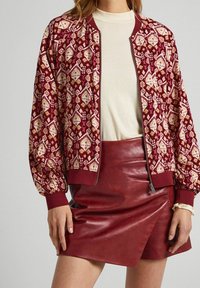 Burgundská bomber bunda s vzorem, zapínáním na zip a žebrovanými manžetami, v kombinaci s lesklou červenou mini sukní z umělé kůže. Hladká textura, vícevrstvý design.