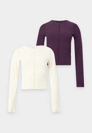 Jeune femme debout portant un haut violet ajusté à manches longues, une jupe en jean bleue et des bottes noires montantes sur fond blanc.