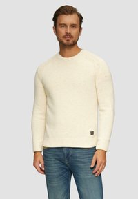 Cremfarbener Strickpullover mit strukturiertem Finish, geripptem Kragen und Bündchen sowie einem kleinen Logo-Patch nahe dem Saum. Kombiniert mit blauen Jeans.