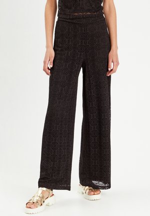 Schwarze Spitzen-Hosen mit weitem Bein, die ein kreisförmiges Muster und einen hohen Bund aufweisen, kombiniert mit goldenen, schnürbaren Sandalen und einem strukturierten Oberteil.