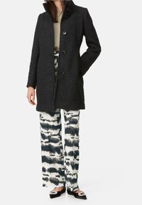 Sort tekstureret frakke med høj krave og knaplukning, kombineret med mønstrede bukser med vide ben i nuancer af blå og hvid.