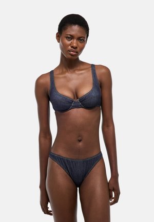 Femme portant un ensemble bikini en denim bleu foncé, debout devant un fond gris clair uni, face à l'avant avec une expression neutre.