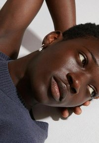 Gros plan sur un visage avec des cheveux courts et foncés, portant une boucle d'oreille en argent. La peau a une texture lisse et l'arrière-plan est de couleur claire.