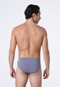 Uomo con capelli scuri di spalle, che indossa slip da mare grigio chiaro su uno sfondo bianco uniforme.