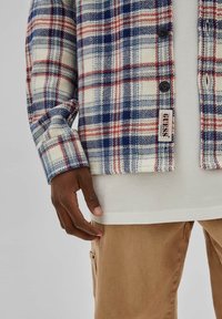 Modello maschile che indossa una camicia a quadri blu e rossa con bottoni neri, una maglietta bianca sotto e pantaloni beige, con la mano rilassata al fianco.