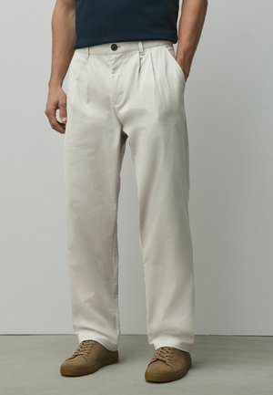 Mann trägt elfenbeinfarbene plissierte Hose mit seitlichen Taschen, braune Sneaker und ein dunkles, in die Hose gestecktes Hemd, steht vor einer schlichten Wand.