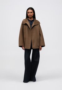 Mujer con una chaqueta oversize marrón de mangas anchas, pantalones oscuros de pierna ancha y una camisa oscura abotonada, de pie frente a un fondo liso.