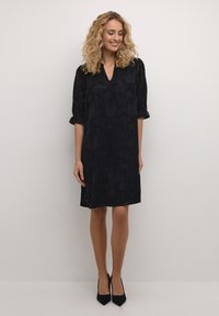 Abito nero con motivo floreale, scollo a V e maniche a sbuffo trasparenti. Il materiale sembra leggero e texturizzato. Indossato con tacchi neri a punta.