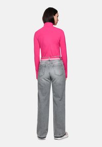 Figurbetontes pinkes Langarmoberteil mit hohem Kragen, kombiniert mit lockeren grauen Jeans mit Gesäßtaschen und einem passendenden Gürtel.