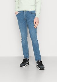 Jeans de denim azul claro com um corte slim, apresentando cinco bolsos e costuras de contraste. Combinados com ténis desportivos pretos e brancos.