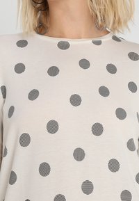 Helles beige Oberteil mit schwarzen gestreiften Pünktchen. Hat einen runden Halsausschnitt und besteht aus einem weichen Stoff. Akzente sind runde Ohrringe.