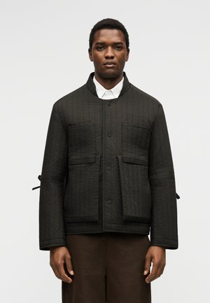 Man wearing black quilted jacket with front pockets and dark brown pants, standing against plain light background.