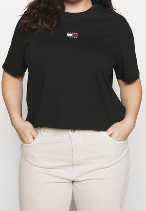 Persoon draagt een zwart Tommy Jeans cropped T-shirt en beige broek met handen in zakken tegen een effen achtergrond.