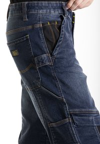 Pantalons de travail bleu foncé en denim, dotés de plusieurs poches, de coutures jaunes et d'un panneau de tissu renforcé sur le côté.