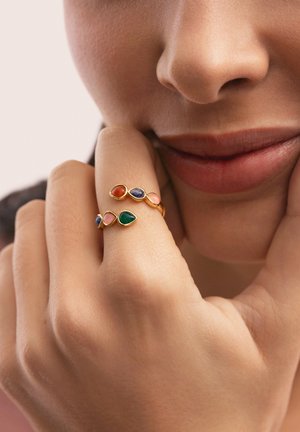 Primo piano di una mano vicino al viso che indossa un anello d'oro con pietre a goccia multicolore sul dito medio.