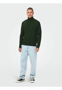 Pulover verde închis cu guler înalt, textură canelată, asortat cu blugi largi de culoare albastră deschisă și adidași negri cu talpă joasă. Fundal neutru.