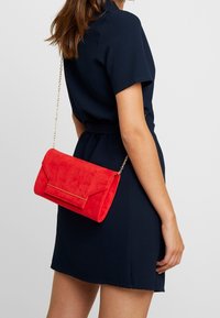 Červená kabelka z umělé semiše s klopovým zapínáním a zlatým řetízkovým popruhem, obdélníkový tvar a hladký povrch.