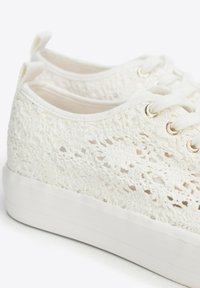 Weiße SpitzenSneaker mit einem strukturierten, gewebten Obermaterial, goldenen Ösen und einer weißen Gummisohle; minimalistisches Design mit Laschen an der Ferse.