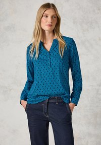 Blusa blu a maniche lunghe con motivo a diamante, colletto con bottoni e vestibilità comoda. Abbinata a jeans in denim scuro.