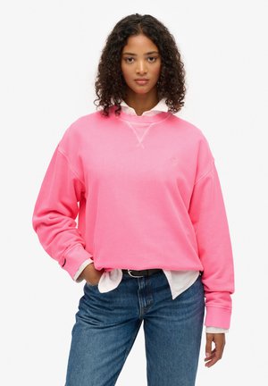 Oversized różowa bluza z ribanem przy mankietach, okrągłym dekoltem i detalem z logotypem. Noszona na białej koszuli z kołnierzykiem i w połączeniu z niebieskimi dżinsami.