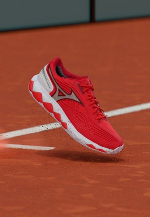 Red athletic shoe with white and red sole, hovering above a clay tennis court near a white boundary line with scattered clay particles.