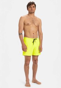 Άντρας που στέκεται ξυπόλητος, φοράει νέον κίτρινα μαγιό με μαύρο κορδόνι, χωρίς πουκάμισο, μπροστά σε απλό λευκό φόντο.