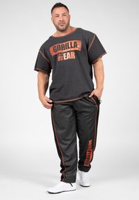 Grijze T-shirt met korte mouwen met "GORILLA WEAR" in oranje, gecombineerd met zwarte broek met oranje zijstreep en logo, en witte schoenen.