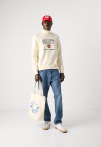 Cremefarbene Sweatjacke mit "GANT"-Logo, blaue Jeans, rote Kappe und eine Canvas-Tasche mit blauem Palmenmuster, weiße Sneakers mit Gummisohlen.