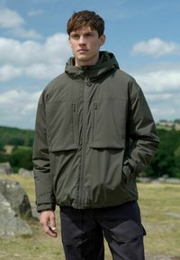 Olivgrön huva/jacka med dragkedjefickor på bröstet och elastiska ärmslut, tillverkad av ett jämnt, isolerat material, som bärs över mörka byxor.