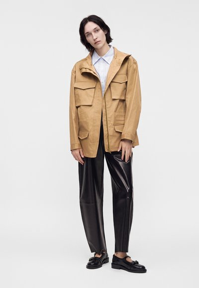 Personne portant une veste beige avec plusieurs poches, une chemise blanche à col, un pantalon en cuir noir à jambes larges et des mocassins noirs sur un fond uni.