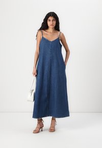 Denim maxi kjole med tynne stropper, V-hals, frontknapper og side lommer. Mørkeblå farge med en glatt tekstur. Kombinert med beige hæler og hvit håndveske.