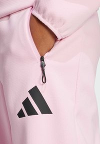 Rosa träningsjacka med en zip-ficka, prydd med en svart Adidas-logga. Materialet verkar texturerat och mjukt, med en avslappnad passform.